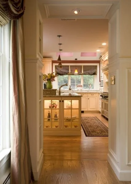 View of a bright kitchen with white cabinets, a large window, and a small island with glass-fronted cabinets. A vase with flowers is on the island, and a patterned rug is on the wooden floor.