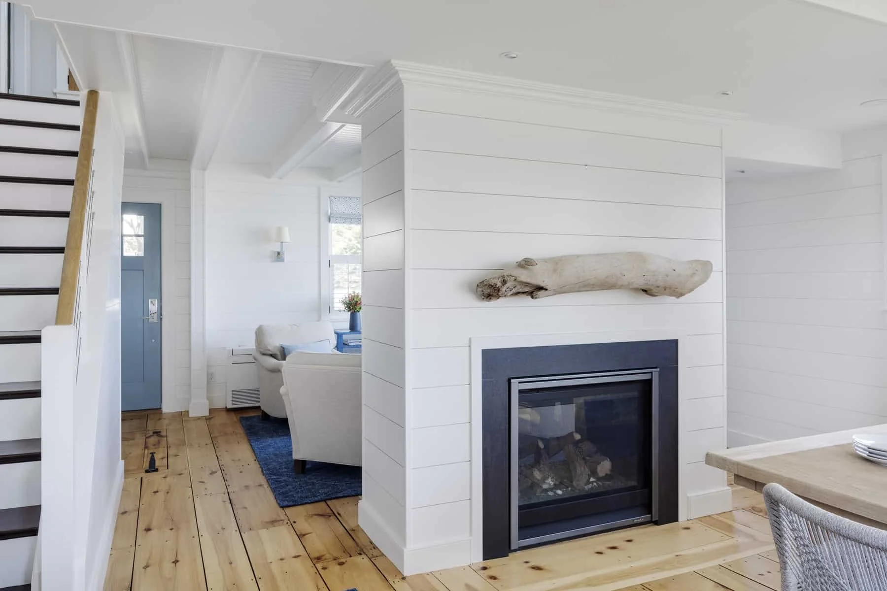 Living room with a white shiplap wall, a silhouette of a driftwood piece above a black-framed fireplace, and a view of a blue door and sitting area with white furniture and a window with a floral arrangement.