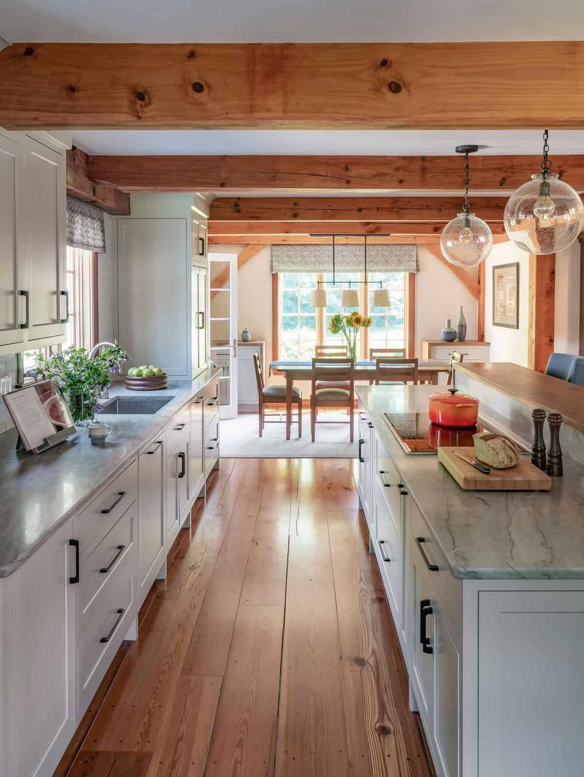 A modern kitchen and dining area with white cabinets, a marble countertop, and wooden beams on the ceiling, featuring a dining table with chairs, hanging pendant lights, and large windows letting in natural light.