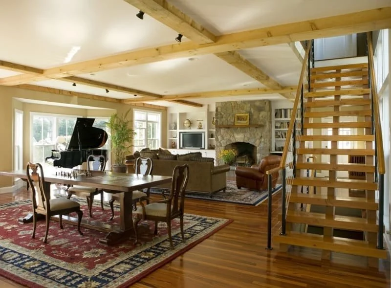 A spacious living room with a dining area, featuring wooden beams, hardwood floors, and large windows; includes a grand piano, a stone fireplace, a TV, couches, and a staircase.