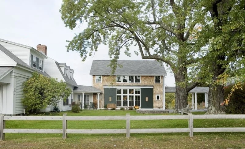 A large house with gray roof and shingles, white and beige siding, large front window, yards with green grass, two large trees, a white fence, and a small porch with chairs.