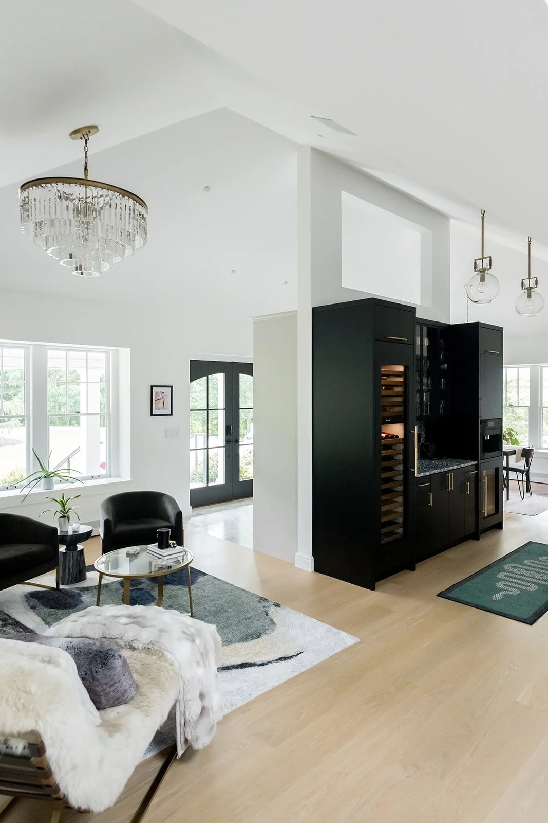 Modern living room with large windows, black furniture, and a black cabinet with wine storage. There are two black chairs, a round coffee table, and a white furry chair. A chandelier hangs from the ceiling, and two glass pendant lights are visible. T