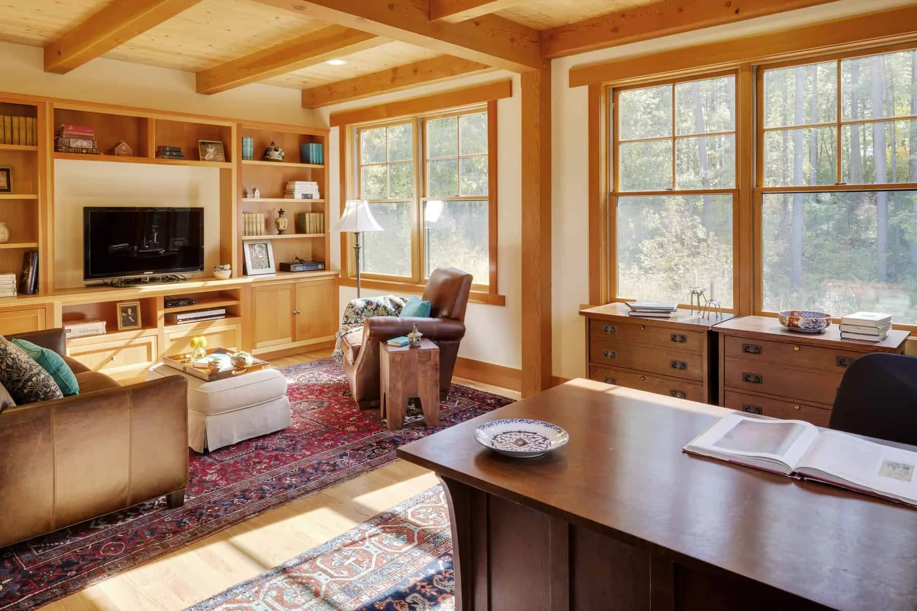 Living room with large windows, wooden ceiling beams, built-in wooden shelves with a television, armchair, sofa, coffee table, and a patterned area rug.