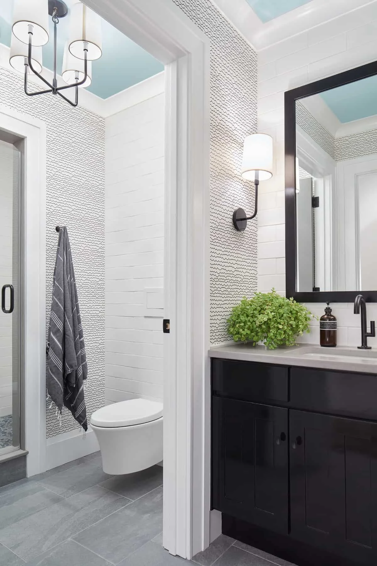 Modern bathroom with white shiplap walls, a black vanity with a beige countertop, a large mirror with a black frame, a wall-mounted light fixture, a potted green plant, and a soap dispenser. A towel hangs on a hook near a shower with glass doors.