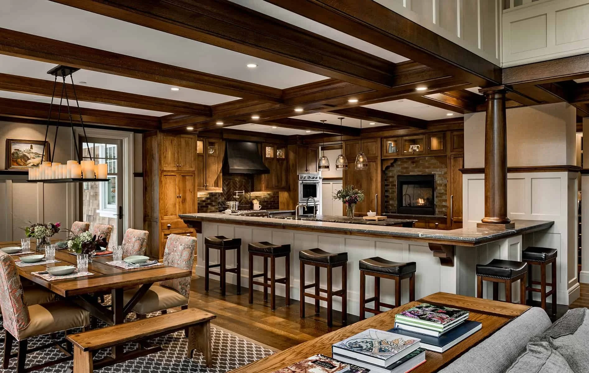 Interior view of a home kitchen and dining area with wooden beams and cabinetry, a fireplace, and a kitchen island with bar stools.