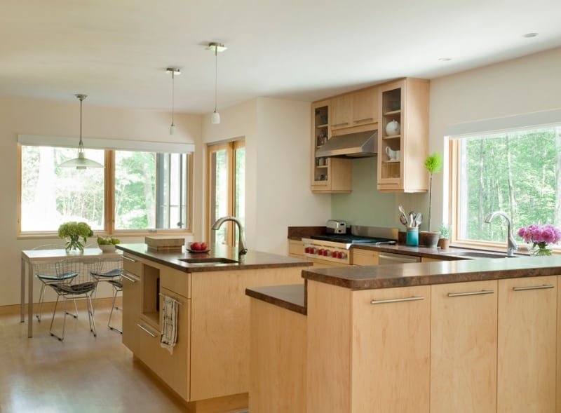 Bright kitchen with wooden cabinets, a central island with a sink, and a dining area with a table and four chairs, large windows showing greenery outside.