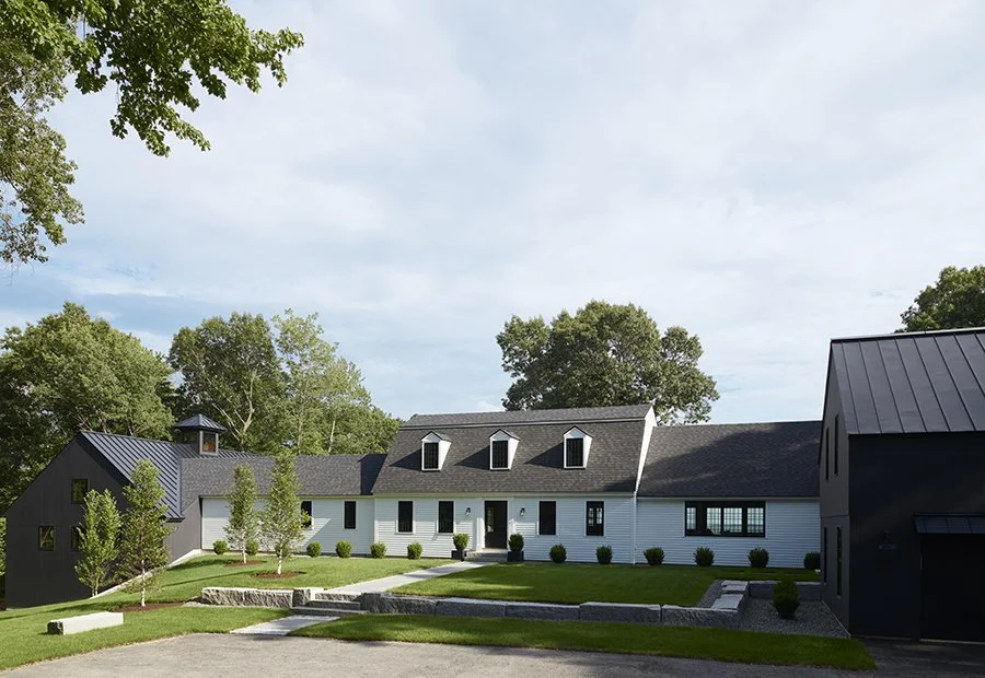 Modern farmhouse with white siding, black roof, and multiple gables, surrounded by green grass, trees, and a gravel driveway under a partly cloudy sky.