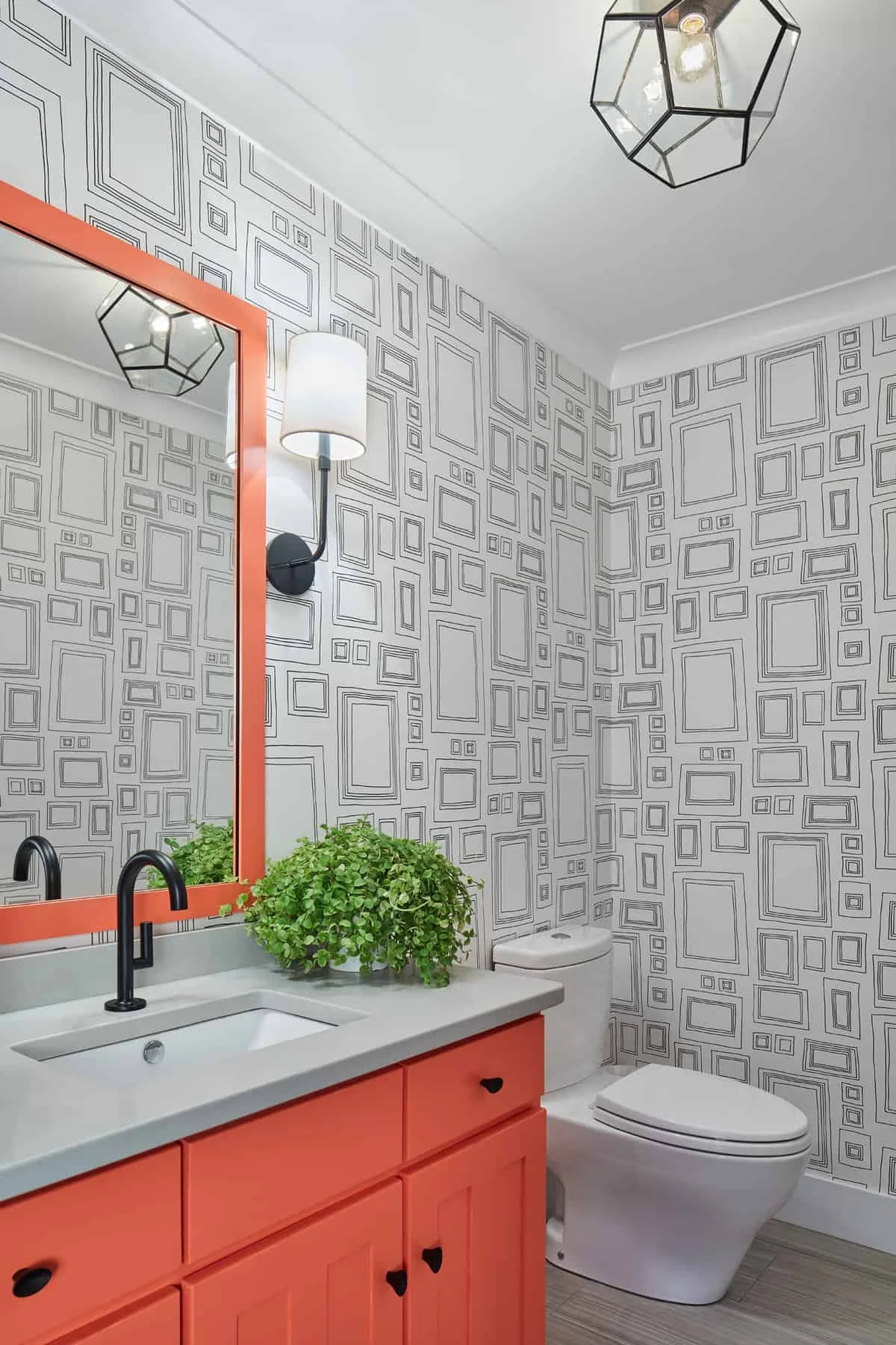 A modern bathroom featuring a vanity with a coral cabinet and a white countertop, a large mirror with an orange frame, a black faucet, a potted green plant on the counter, a white toilet, gray wood-like flooring, patterned wallpaper with geometric sq