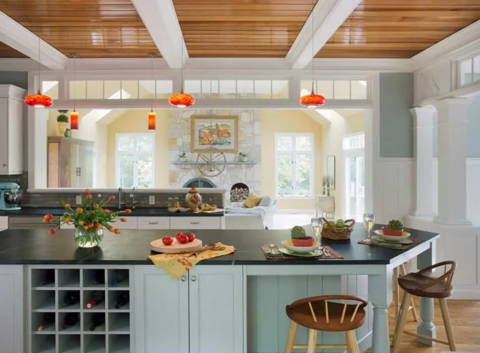 Open-concept kitchen and living area with a wooden ceiling, white walls, and large windows. The kitchen island has a vase of flowers, a cutting board with tomatoes, and place settings with wine glasses and plates. In the background, there's a stone f