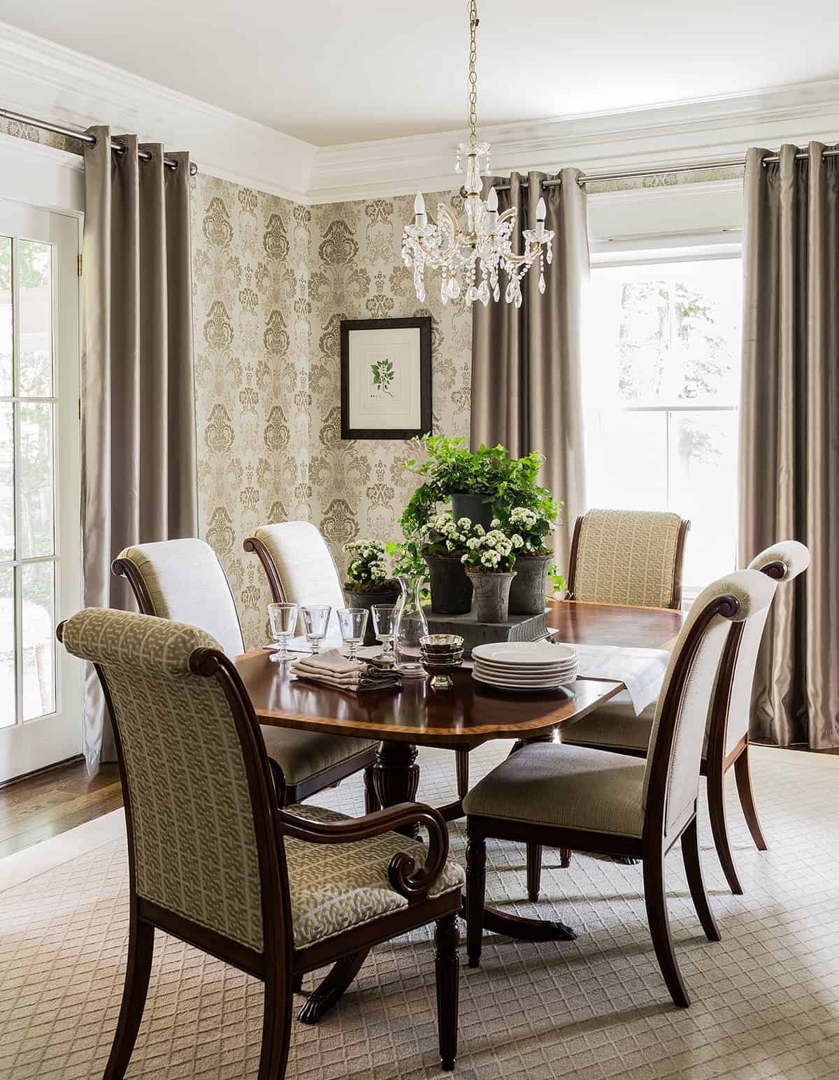 A dining room with a wooden table set with dishes, glasses, and napkins, surrounded by six upholstered chairs. A chandelier hangs above, and potted plants serve as a centerpiece. Large windows with curtains allow natural light, and a framed botanical