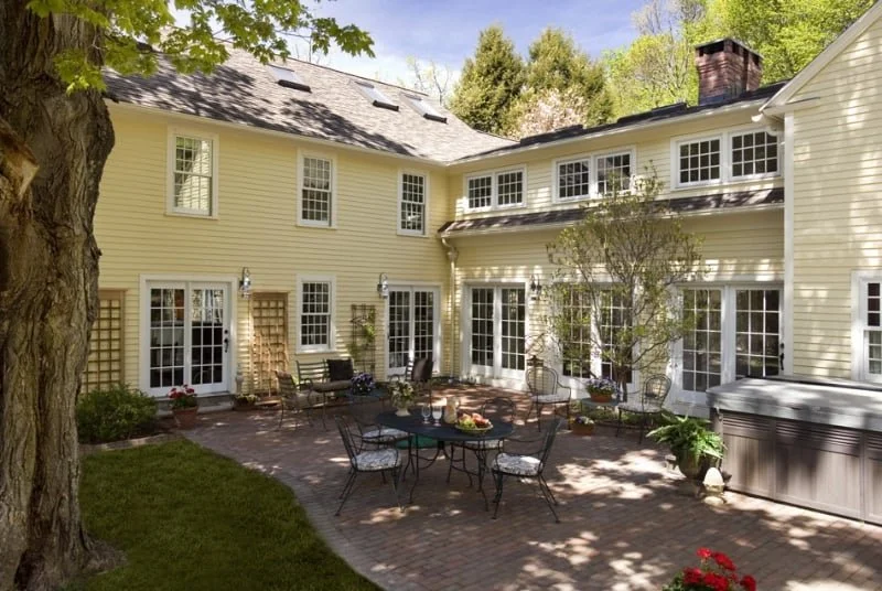 Backyard patio area with outdoor furniture, potted plants, and a yellow house with multiple windows and a brick chimney.