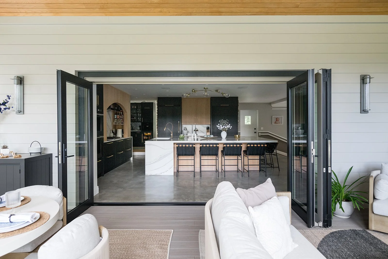 Open black-framed glass doors leading to a modern kitchen and living room with white and black decor, a kitchen island with black bar stools, and a beige sofa.