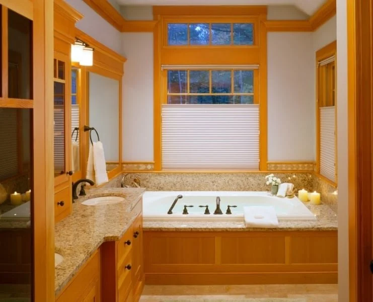 Bathroom with a bathtub, granite countertop, and wooden cabinetry, illuminated by a wall sconce and featuring a window with blinds, candles, flowers, and toiletries.