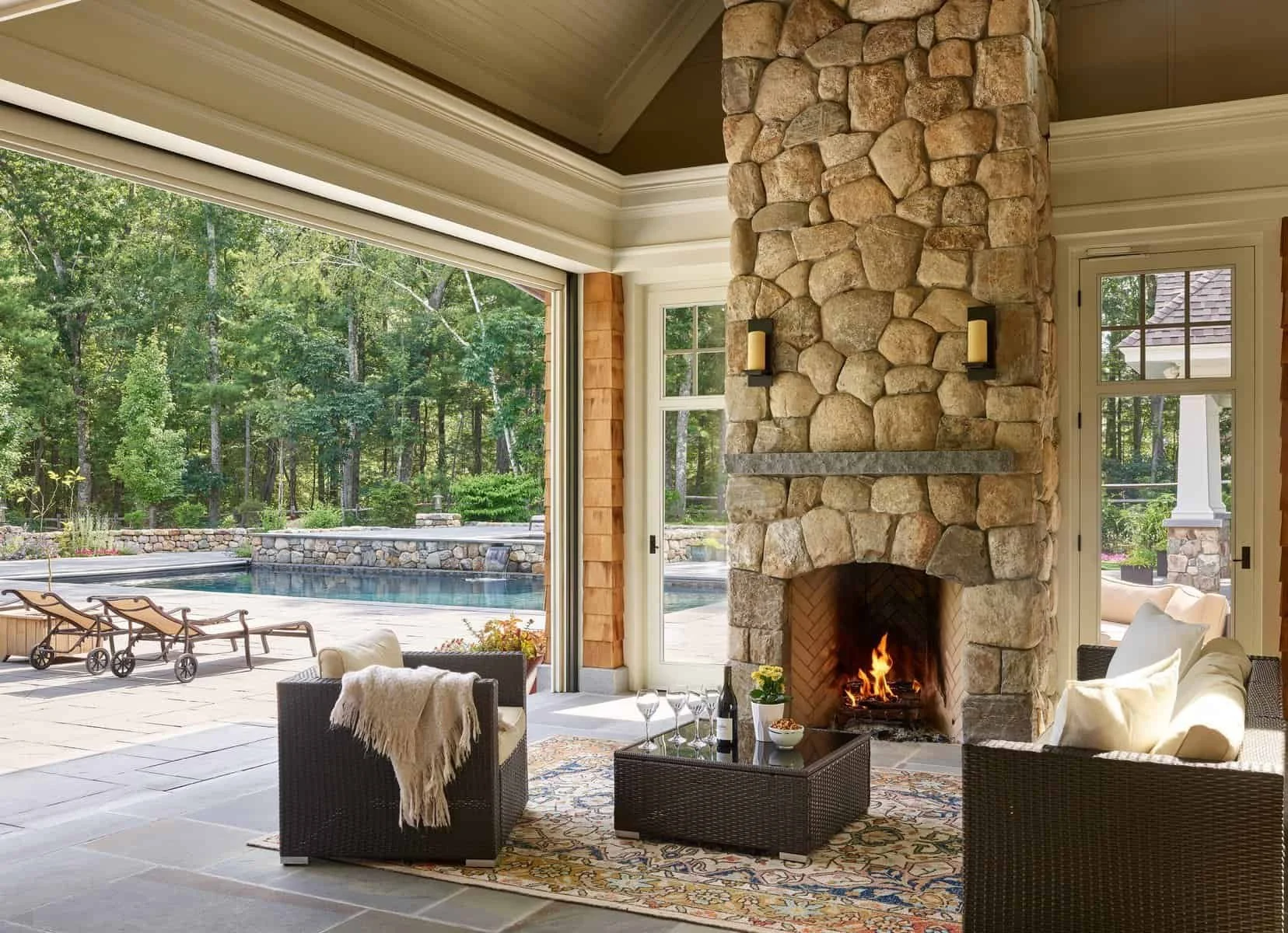 Living room with a stone fireplace, beige sofa with white pillows, wicker chairs, and a table set with wine glasses. Large sliding glass door opens to an outdoor area with a swimming pool, lounge chairs, and a lush forest backdrop.