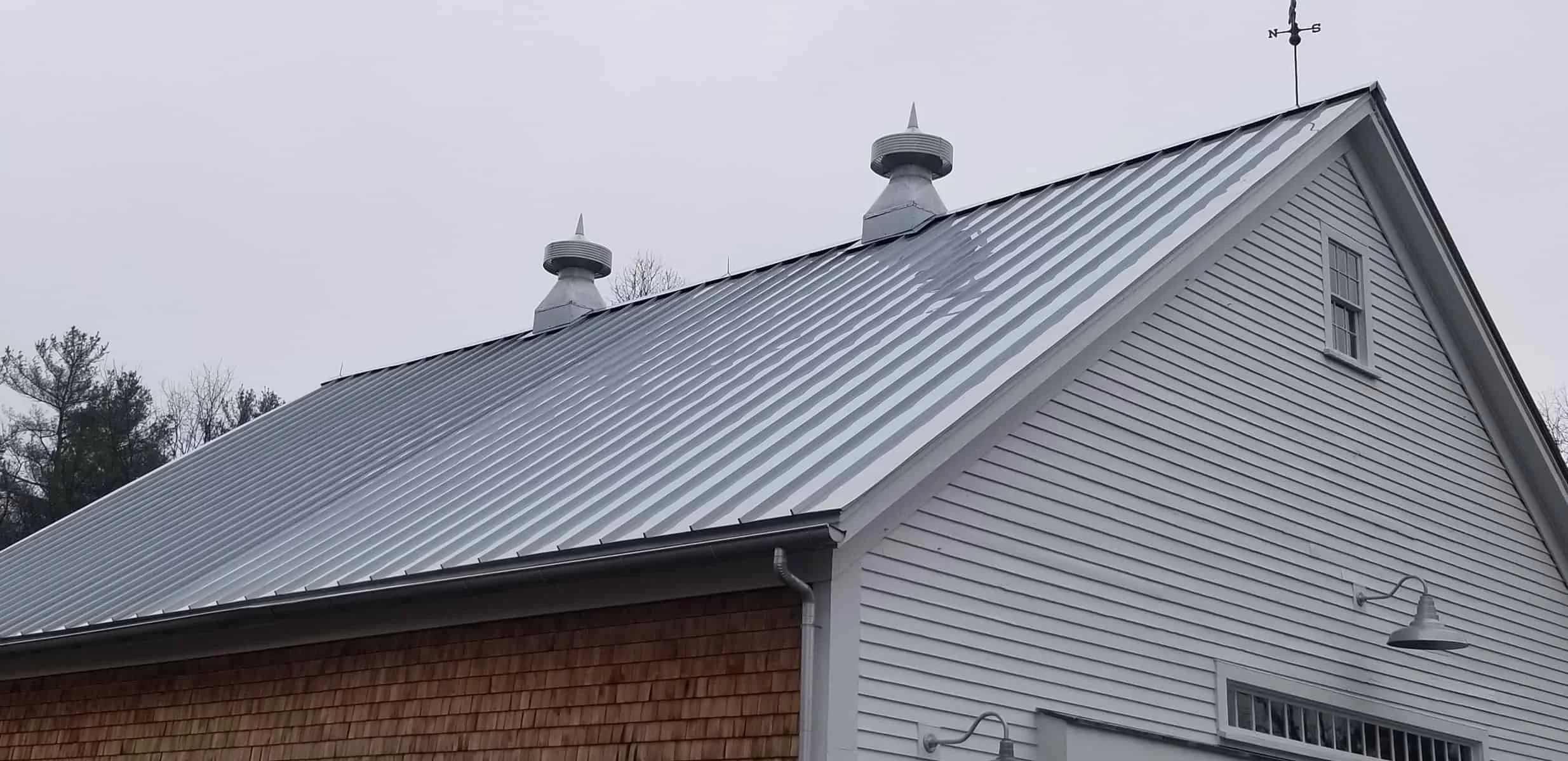 Side view of a house with a metal roof, white siding, and wooden shingles, under an overcast sky, with trees in the background.