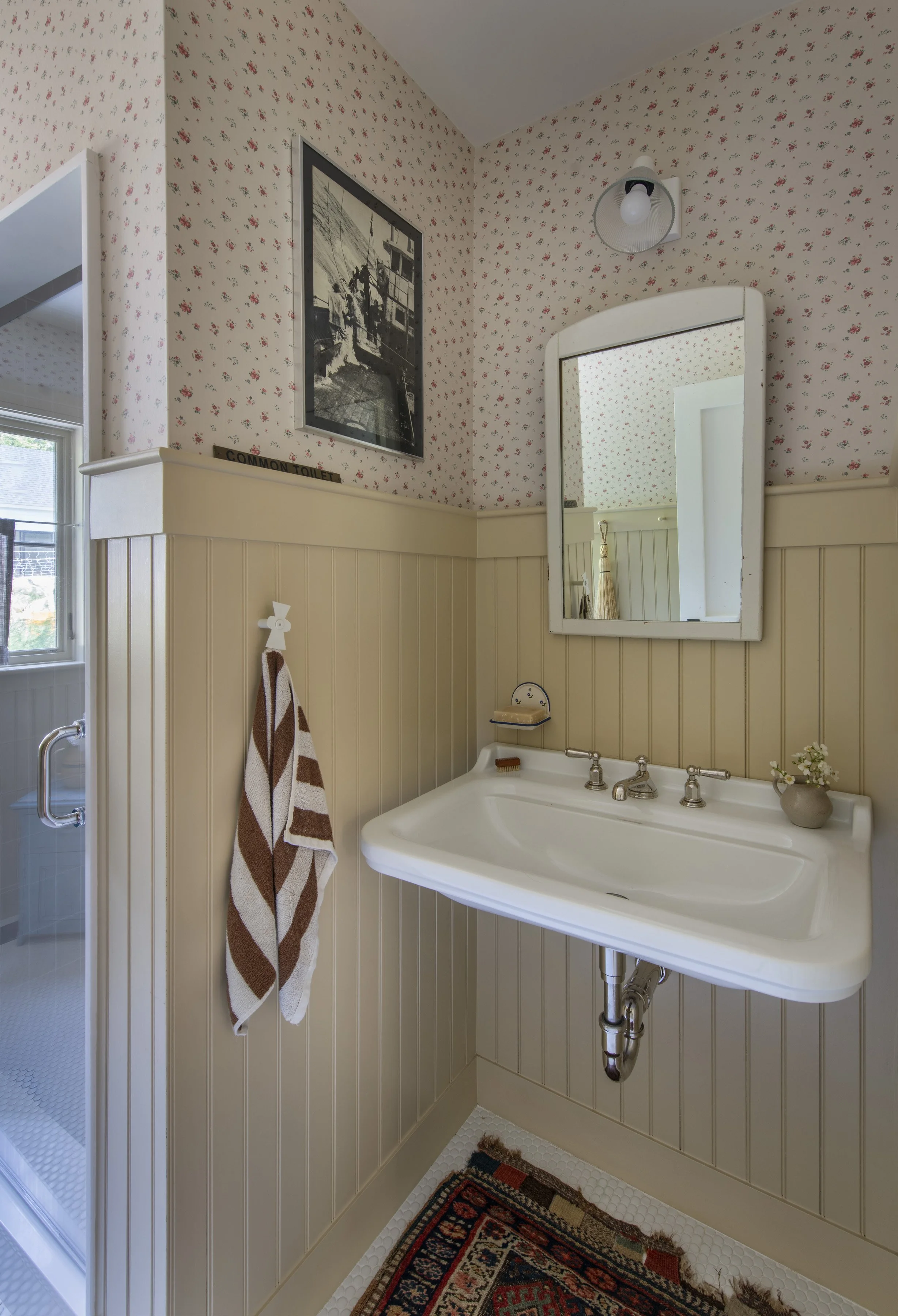 Small bathroom with a vintage sink, a mirror, a towel hanging on a post, floral wallpaper, a mosaic rug, and a wall lamp.