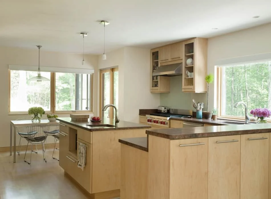 Bright kitchen with light wood cabinets, granite countertops, and large windows showing greenery outside. Features include a kitchen island with a sink, a stove with an exhaust hood, a dining table with chairs, and decorative plants.