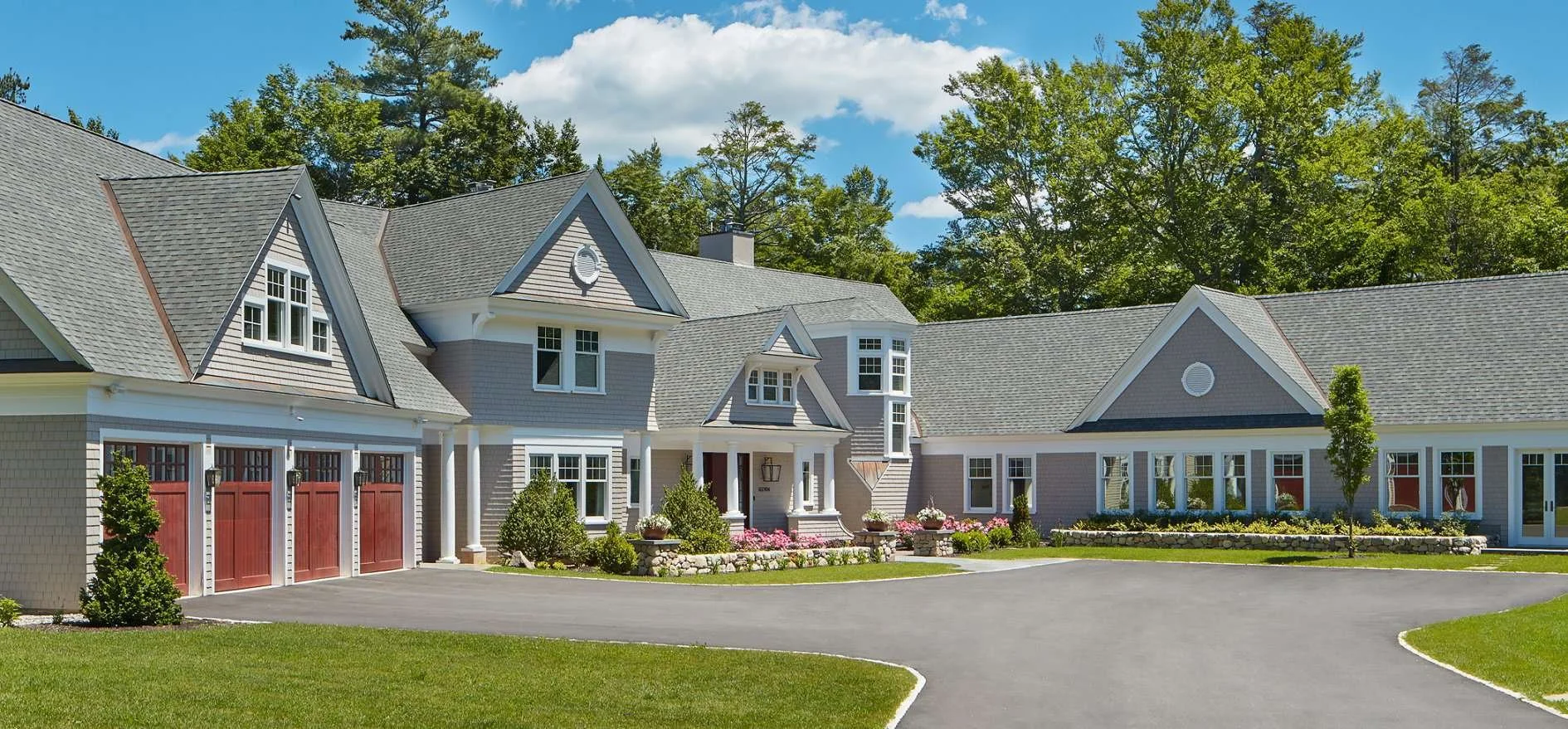 Large suburban house with grey siding, multiple gables, red garage doors, well-maintained garden, and a curved driveway, surrounded by green trees.