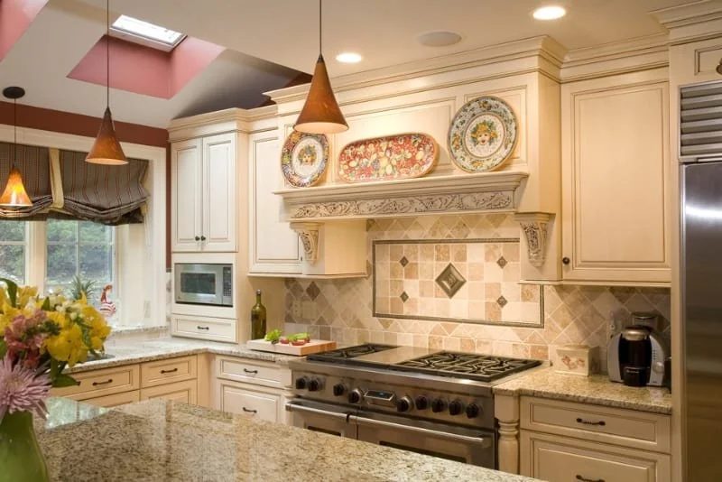 A spacious kitchen with cream-colored cabinets and a granite countertop island. There are decorative plates on the wall above the stove, which has a tiled backsplash with diamond-shaped accents. Pendant lights hang over the island, and there is a sma