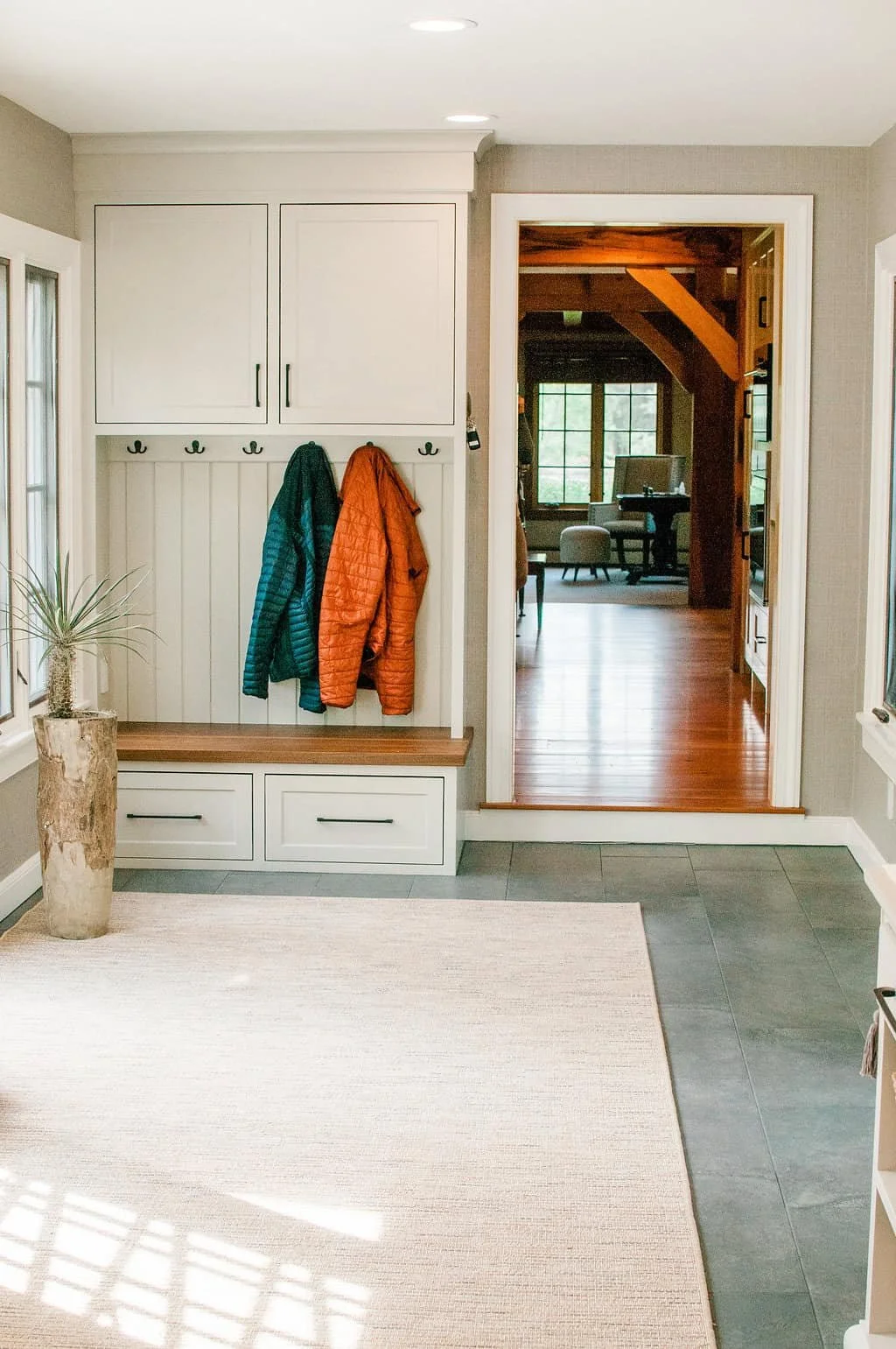 Entryway with white built-in storage, coat hooks with two jackets hanging, a beige rug, a potted plant in a wooden planter, and a doorway leading to a room with wooden floors and large windows.