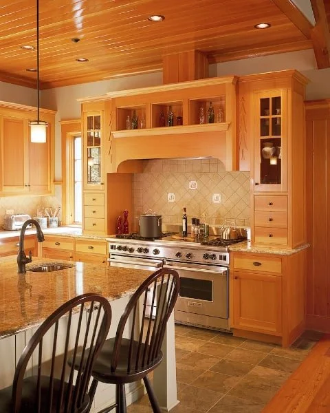 Wooden kitchen with a granite countertop island, a stainless steel stove, and wooden cabinets. There are bar stools at the island, a window behind the sink, and a wooden ceiling.
