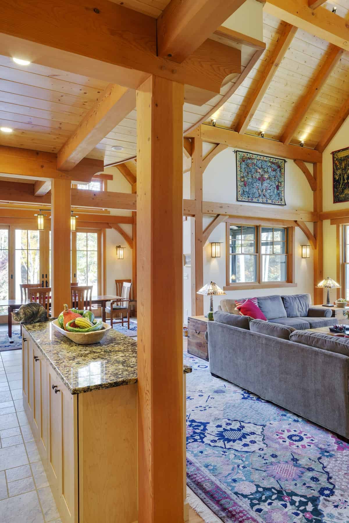 Open-concept living room and dining area with wooden beams, large windows, a gray sofa, and a colorful rug. Kitchen island with a bowl of fruit and a cat. Decor includes wall hangings and table lamps.