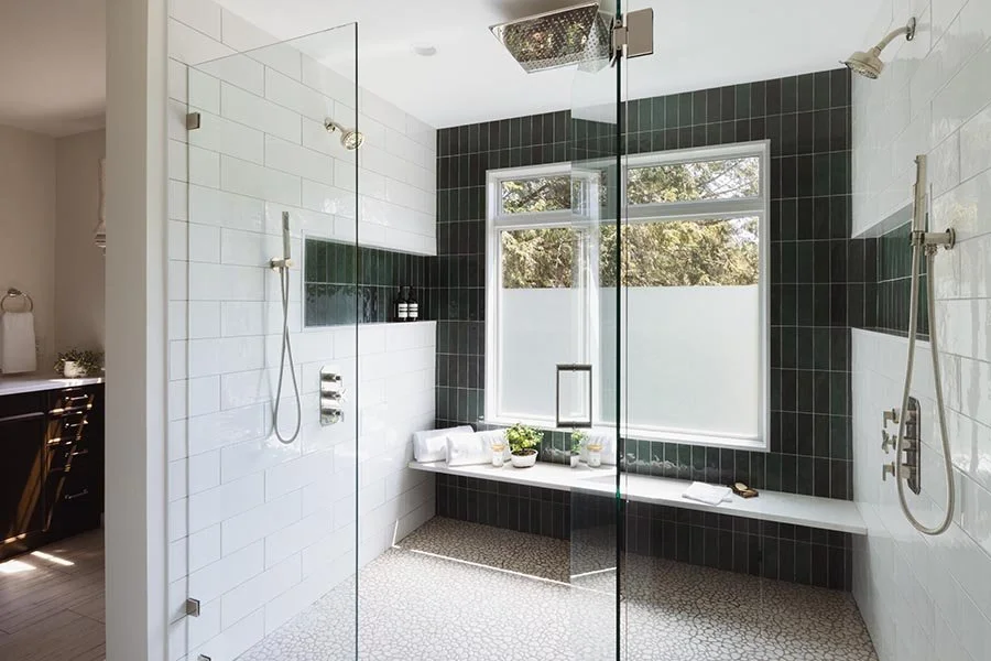 Modern bathroom with a walk-in shower, black tiled wall, white tiled wall, large window with frosted lower section, and a built-in seating area with plants and towels.