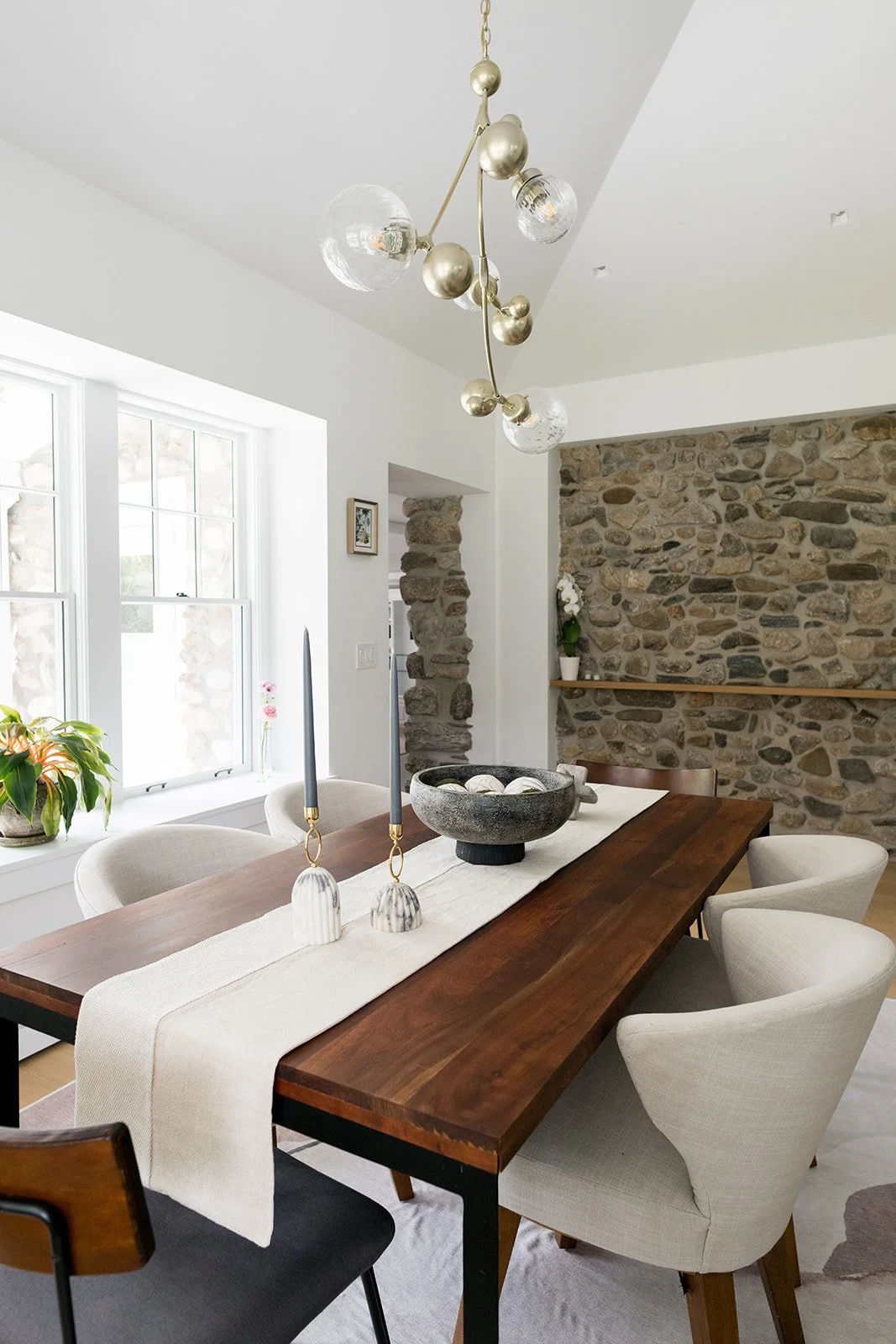 A dining room with a wooden table, beige upholstered chairs, a stone accent wall, and a modern chandelier with gold and glass globes, near large windows with houseplants.