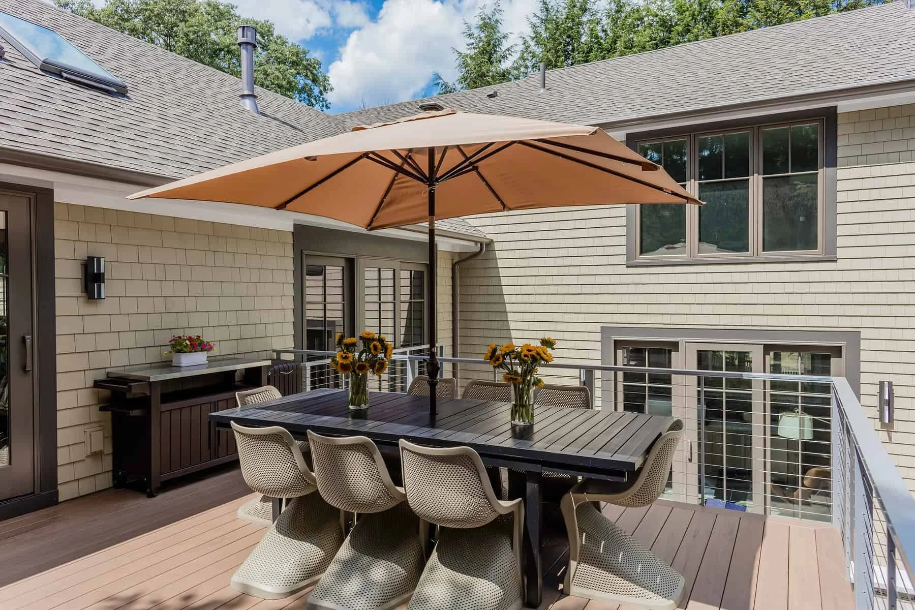 Outdoor patio with a dining table, chairs, sunflowers, a large umbrella, and a view of the house with large windows and beige siding.