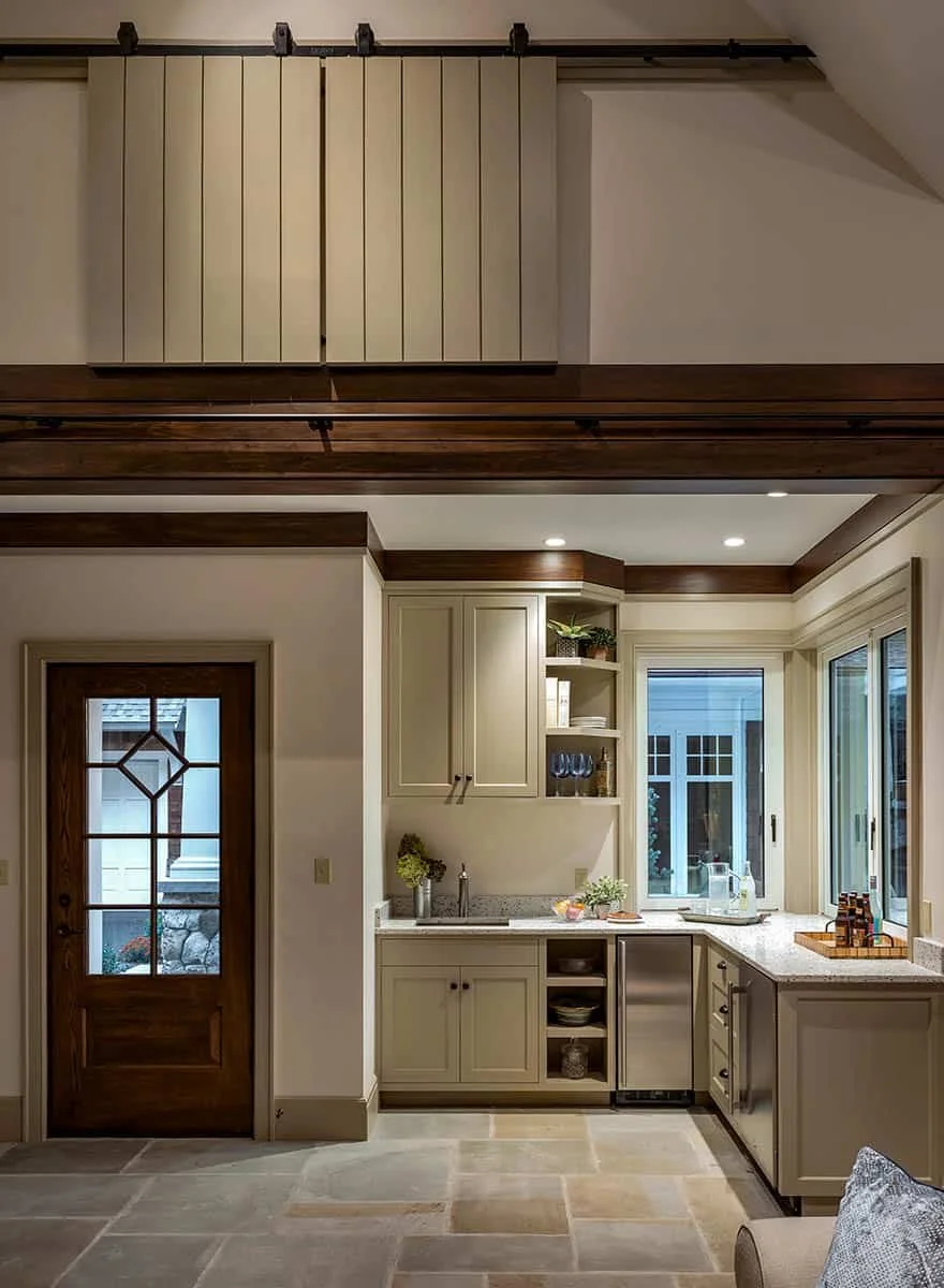 A cozy kitchen with white cabinetry, granite countertops, and large windows, adjacent to a wooden door with glass panels, under a ceiling with recessed lighting and dark wood trim.