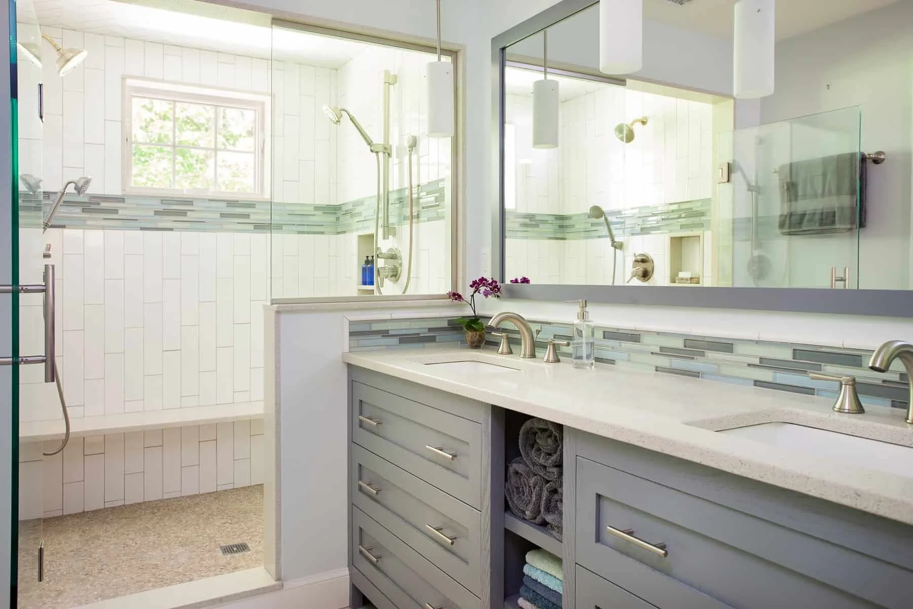 Modern bathroom with light gray vanity, white countertop, large mirror, and glass-enclosed shower with white tile and a decorative gray-green tile border. The shower has a window, showerhead, and built-in niche with toiletries. The vanity has rolled 