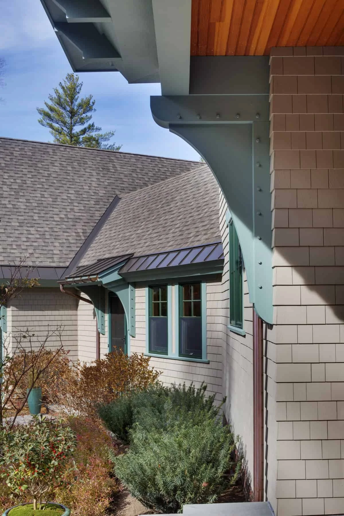 Close-up of a house's architectural details, including a roof with shingles, a curved teal soffit with decorative brackets, and a brick exterior with teal window trim, surrounded by shrubs and trees.