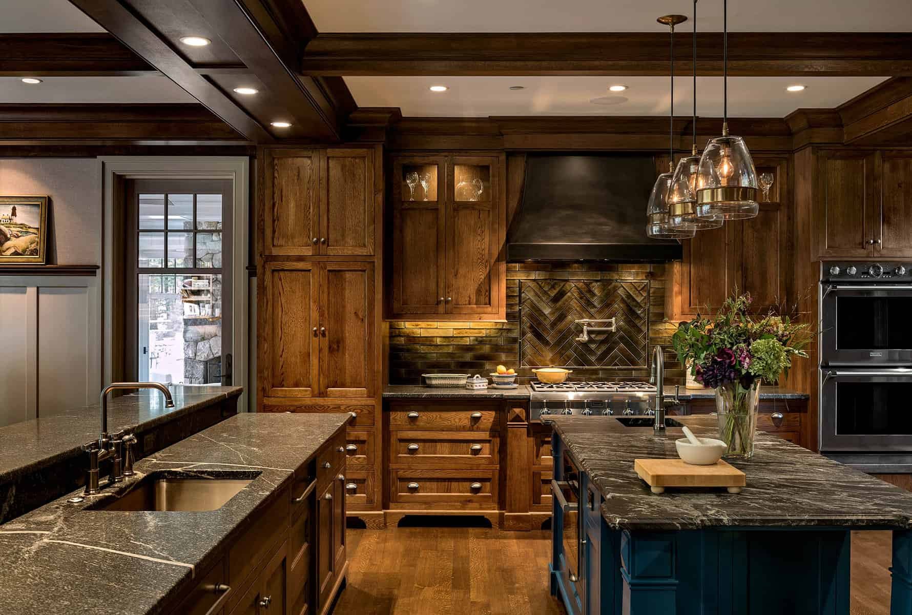 A kitchen with dark wood cabinets, black countertops, a stainless steel stove and oven, a black range hood, a vase with flowers on the island, pendant lights, and a glass door leading outside.