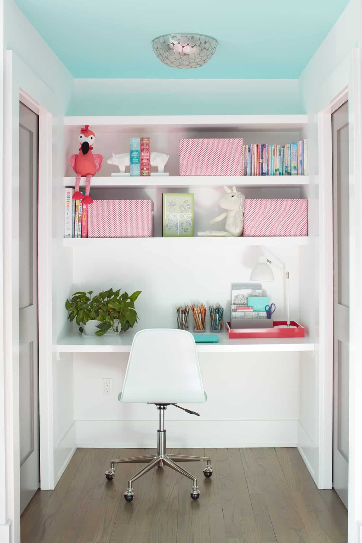 A built-in white desk area with shelves, a white chair, a green potted plant, and office supplies in red and clear containers. The shelving holds books, pink polka-dot boxes, and stuffed animals, including a pink flamingo and a white horse. The ceili