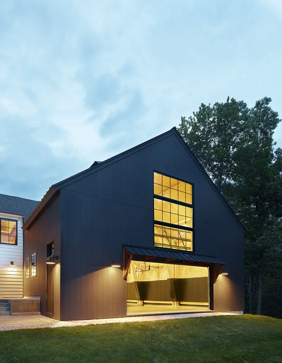 A modern black garage building with large upstairs windows, open garage door revealing an indoor basketball court, illuminated by exterior lights at dusk, with a grass lawn in the foreground and trees in the background.