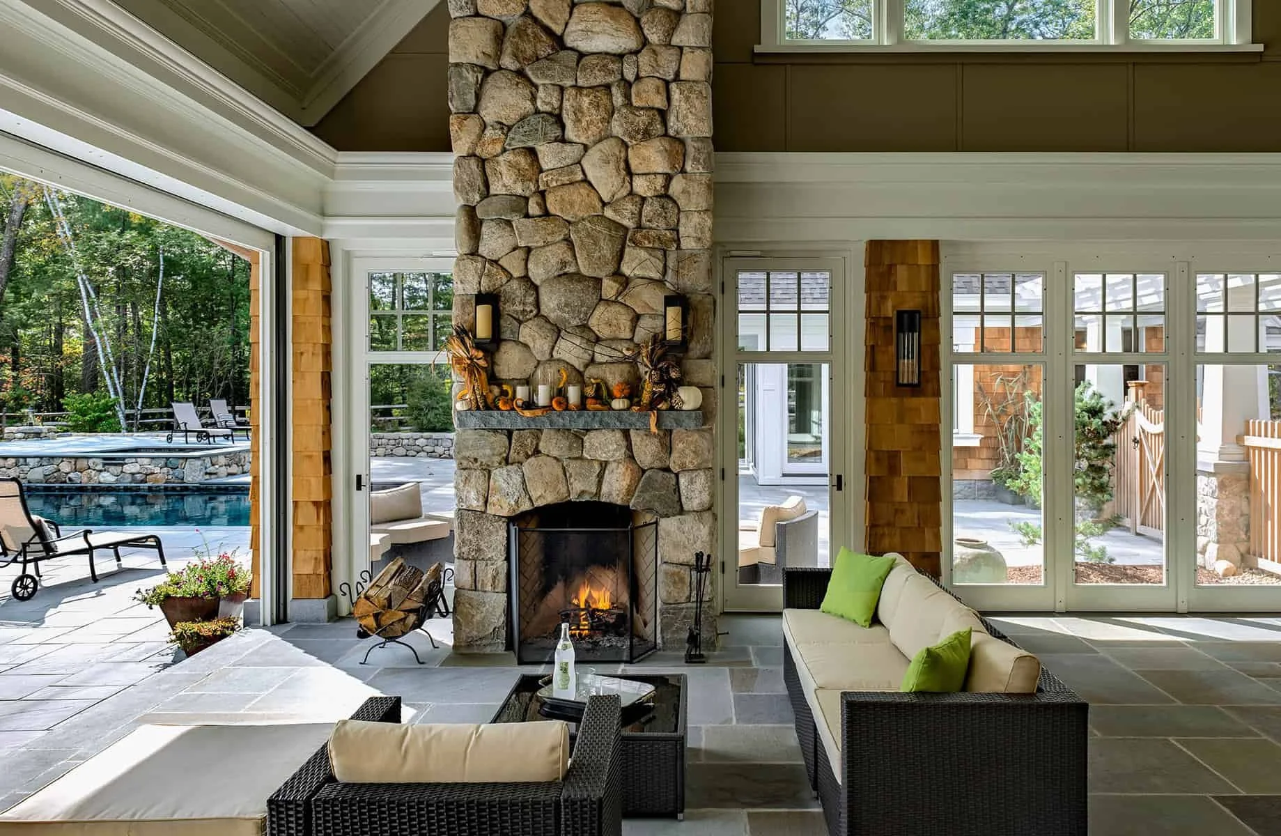 Living room with a stone fireplace, hardwood floors, and large windows overlooking a backyard with a pool and trees.