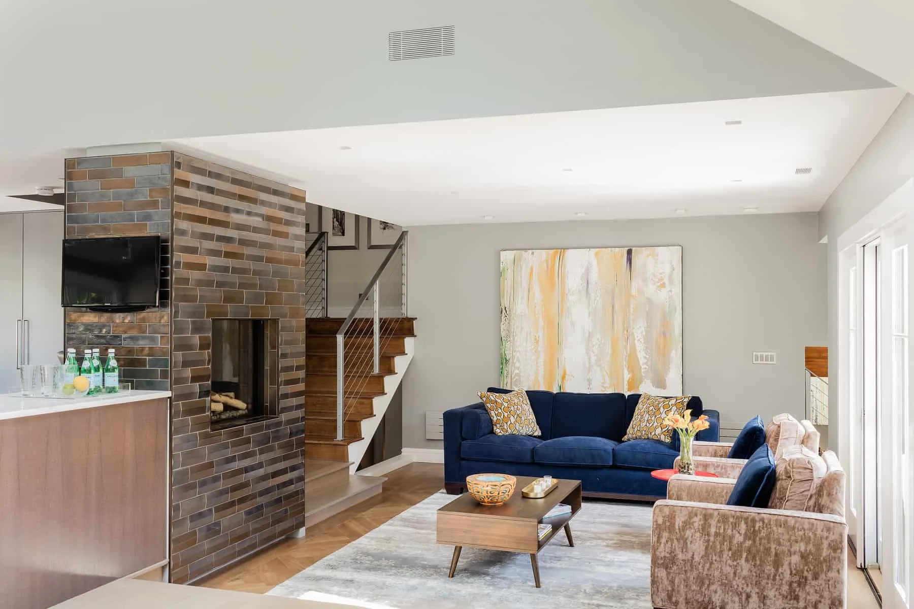 Modern living room with blue sofa and patterned armchairs, abstract wall art, wooden staircase, brick fireplace, and a portion of kitchen with bottles on counter.