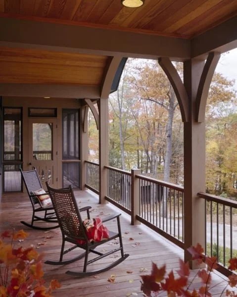 A wooden porch with rocking chairs and fall leaves, overlooking a forest with colorful autumn trees.
