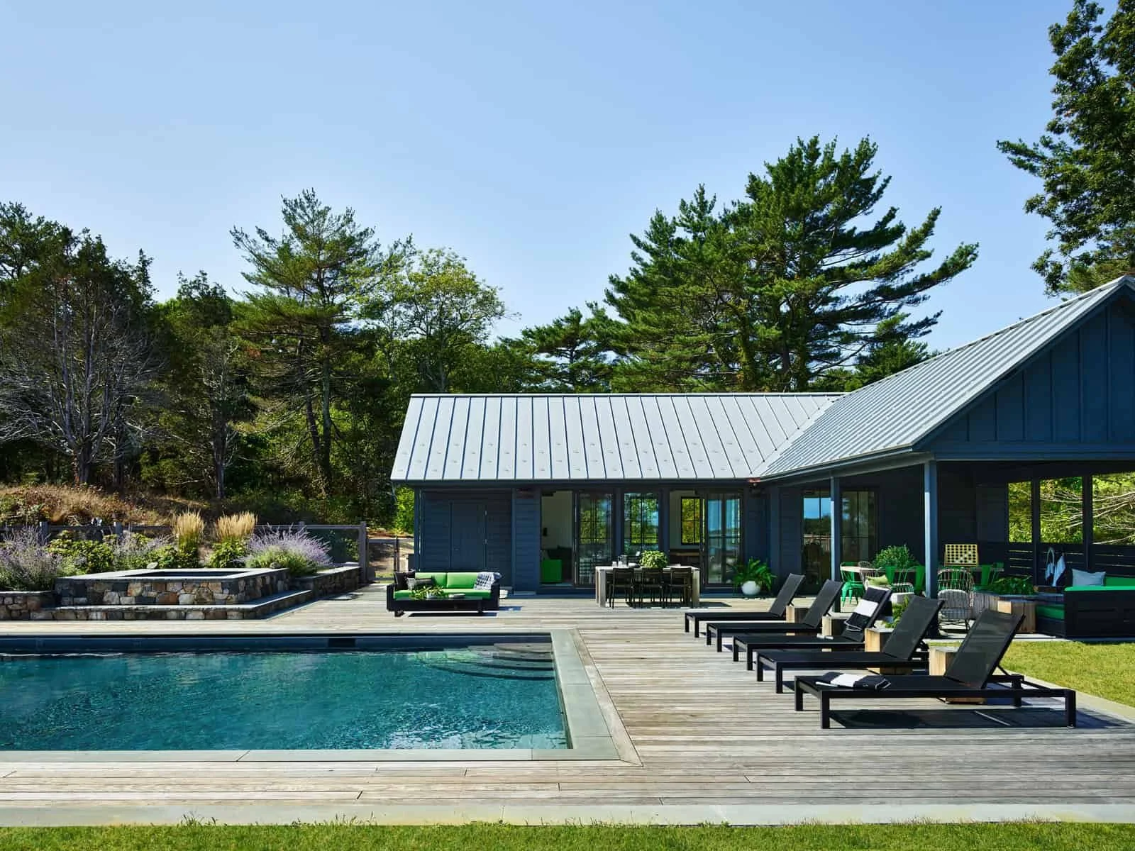 A backyard with a swimming pool, outdoor furniture including lounge chairs, a dining table, and sofa, a grey house with large windows and a metal roof, surrounding trees and landscaped plants.