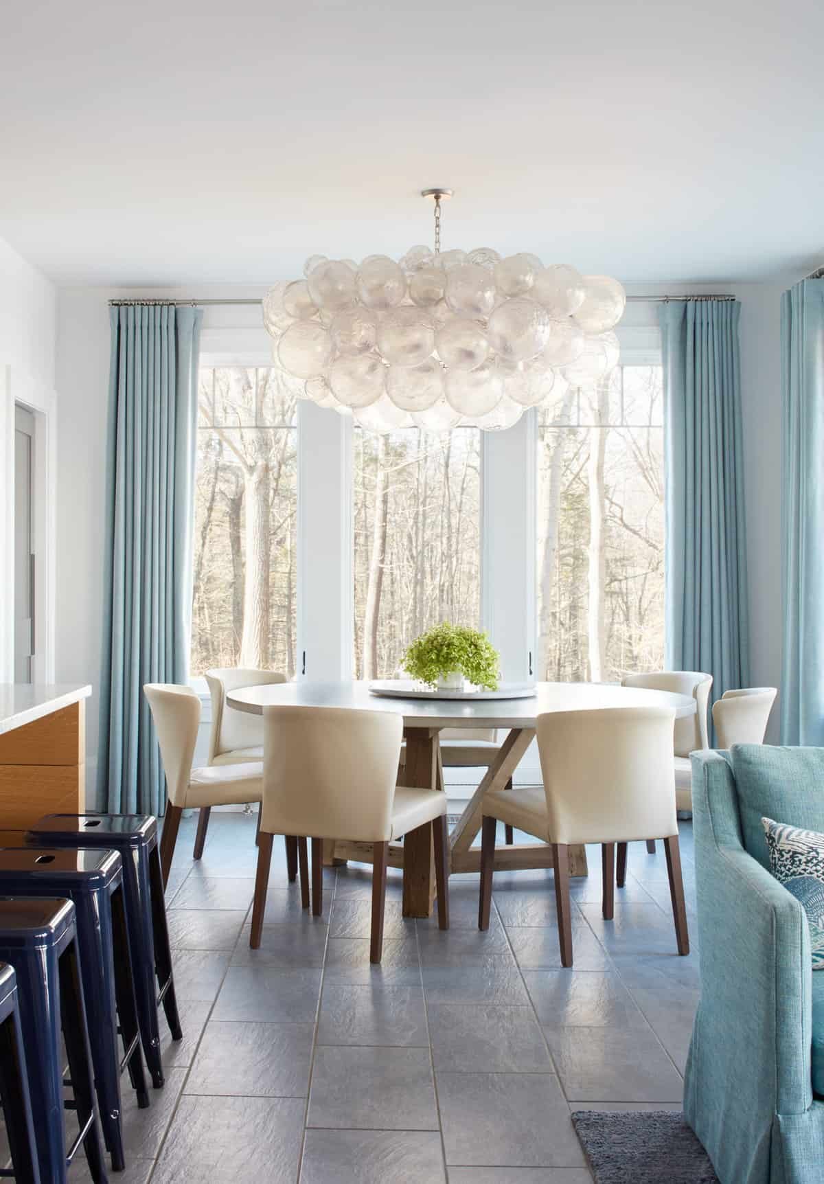 A dining room with a round wooden table, beige chairs, a white chandelier made of glass bubbles, large windows with light blue curtains, and a view of trees outside.