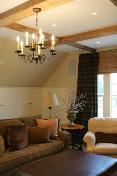 Living room with a beige couch, white armchair, wooden side table with a vase of flowers, and window with plaid curtains. Chandelier and ceiling lights illuminate the room.