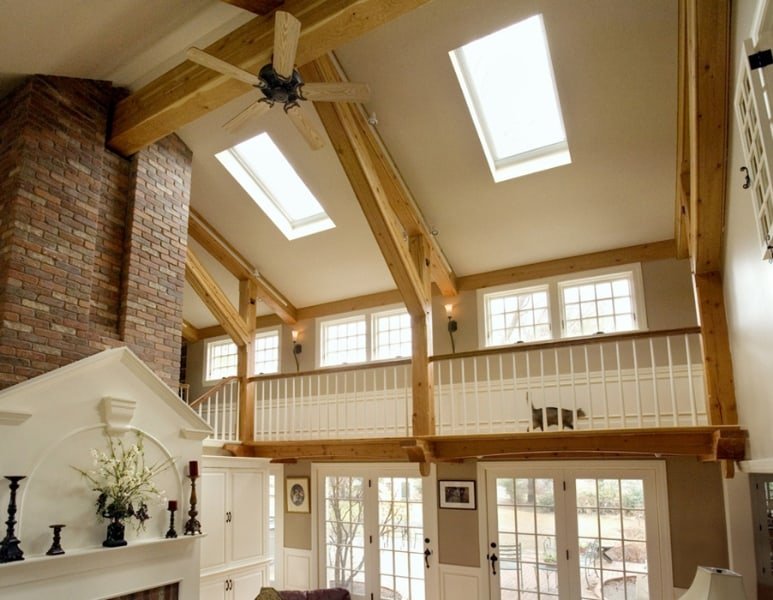 Interior of a living room with high vaulted ceiling featuring exposed wooden beams, skylights, and large windows, with a brick chimney and a white fireplace mantel decorated with candle holders and flowers.