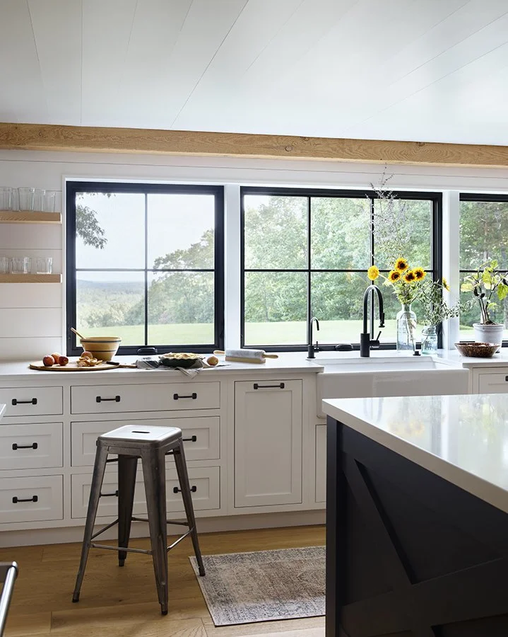 Modern kitchen with white cabinets, black handles, and a large window with a scenic outdoor view. A stool is in front of the cabinets, and a countertop has a bowl, baking ingredients, and a rolling pin. There are sunflowers in a vase, and potted plan