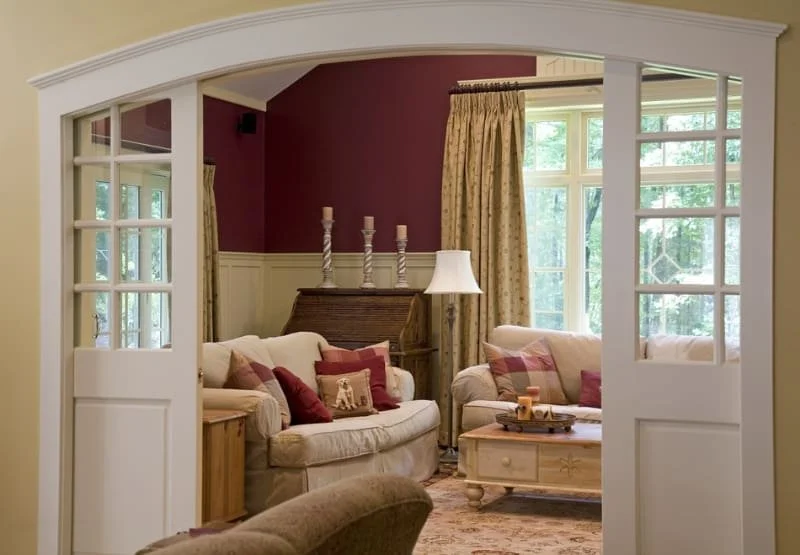 Living room with cream sofas, burgundy wall, large windows with yellow curtains, coffee table with tray, and decorative candlesticks.