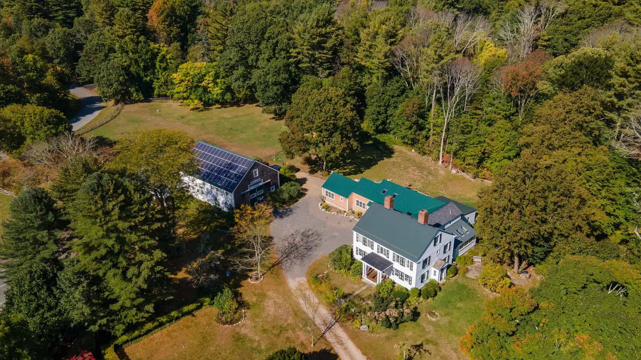Aerial view of a rural property with a large house, a barn with solar panels, and a wooded yard.