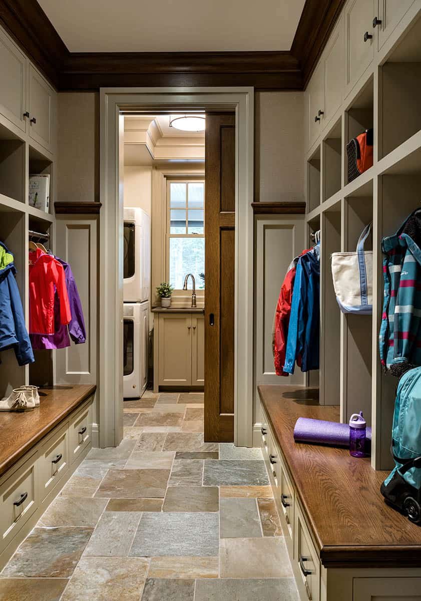 A spacious mudroom or laundry room with built-in cubbies and hooks for coats and outdoor gear, a wooden countertop on both sides, a purple yoga mat and water bottle, and a laundry area with a washer and dryer at the end, small sink, and a window.