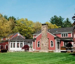 A large red house with multiple sections, a stone chimney, and a well-maintained green lawn in front, surrounded by trees.