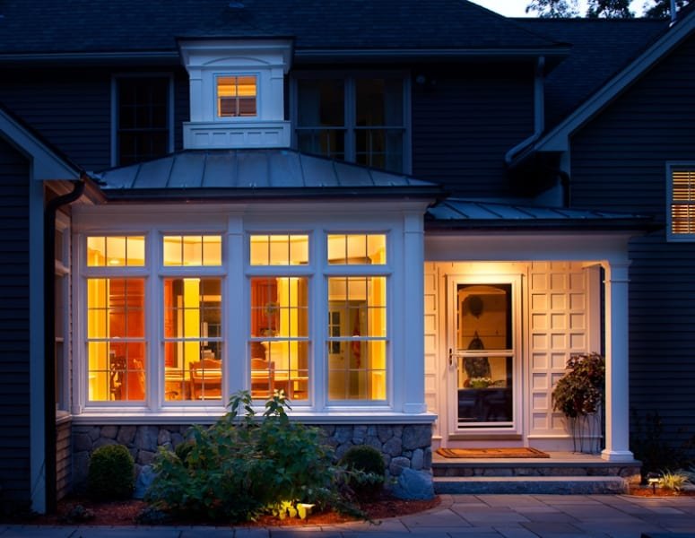 Well-lit house with a bay window and front door at dusk, surrounded by landscape plants and stone foundation.