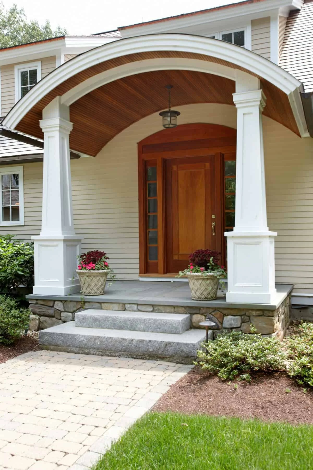 Front porch of a house with stone steps leading to a wooden front door, flanked by two large white columns supporting a curved wooden porch roof, with potted flowers on each side and landscaped garden with bushes and grass.