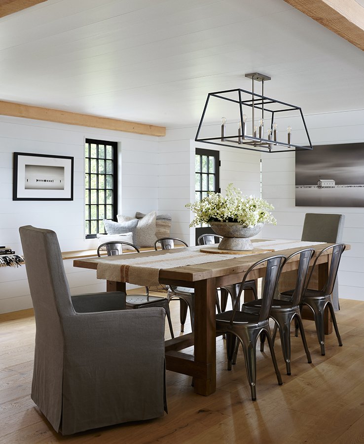 A modern dining room with a large wooden table, six metal chairs, and two upholstered chairs. A centerpiece with white flowers is on the table. The room has white shiplap walls, two black-framed windows, and wall art. A geometric chandelier hangs abo